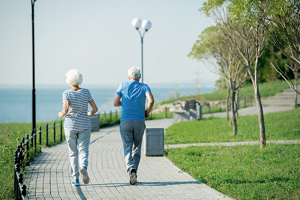 Back view portrait of active senior couple running on park lane along sea shore outdoors , copy space